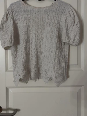 Vit spetsblus från Zara - Supersöt vit blus från Zara med korta puffärmar och spetsdetaljer längs nederkanten. Blusen har ett vågigt strukturmönster och rund halsringning. Perfekt för dig som gillar romantiska och feminina toppar. Den är i storlek 164 13-14 år och är användsa fåtal gången men som ny