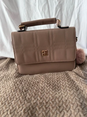 Beige/brun handväska  - Stilren beige handväska i fyrkantig modell med rutmönster på framsidan och guldfärgad detalj. Väskan har ett kort handtag upptill och en fluffig beige nyckelring som är avtagbar. Tillverkad i slätt konstläder med guldfärgade metalldetaljer.