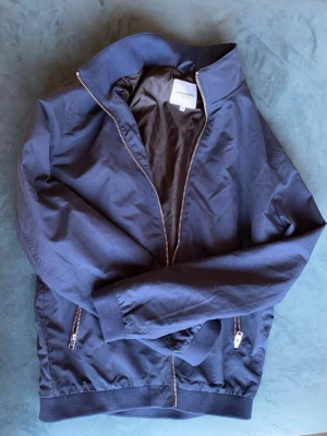 Mörkblå bomberjacka Jack & Jones - Snygg mörkblå bomberjacka från Jack & Jones med dragkedja framtill, ribbade muddar och två fickor med dragkedja. Jackan har klassisk bomberkrage och är tillverkad i ett lätt material som passar perfekt till streetwear-stilen.