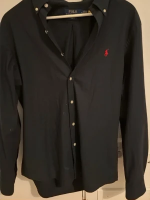 Svart skjorta från Polo Ralph Lauren - Klassisk svart skjorta från Polo Ralph Lauren med röd broderad logga på bröstet. Skjortan har lång ärm, knappar framtill och button-down krage. Perfekt till både jeans och chinos för en clean look. Den är äkta köpt från care of carl 