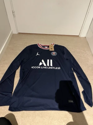 PSG långärmad matchtröja navy - Mörkblå långärmad matchtröja från PSG med vit logga och texten 'Accor Live Limitless' på bröstet. Röd, vit och blå randig krage, PSG-märke på vänster bröst och Jordan-logga på höger. Tillverkad i lätt funktionsmaterial, perfekt för fotboll.