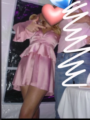 Snygg rosa klänning med volang - Säljer en superfin rosa kortklänning i satinlook med v-ringning, volangdetaljer i midjan och vid ärmslut samt långa ärmar. Klänningen har en omlottkänsla och stängs med dragkedja bak. Perfekt för PinkParty!🙈💕