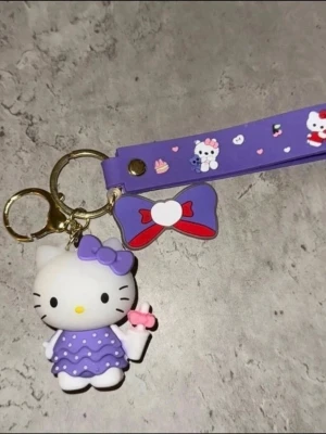 Lila Hello Kitty nyckelring - Söt nyckelring med Hello Kitty-figur i lila klänning och matchande rosett. Nyckelringen har även ett lila band med tryck av Hello Kitty, cupcakes och hjärtan samt en dekorativ rosett i lila och rött. Perfekt att hänga på väskan eller nycklarna.