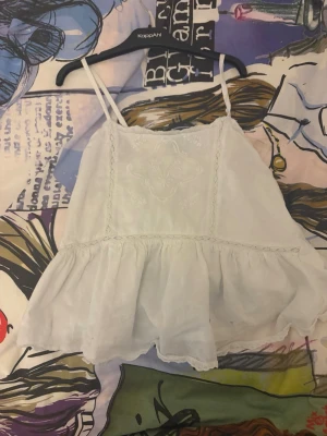 Vit volangig linnetopp från Gina  - Söt vit linnetopp med tunna axelband och volangkant nertill. Toppen har broderade detaljer framtill och är tillverkad i ett lätt och luftigt material, perfekt för varma dagar. Perfekt nu till sommaren 