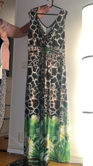 Långklänning med djurmönster och gröna detaljer - Snygg långklänning med giraffmönster i brunt och svart upptill och gröna palmblad samt vita blommor nertill. Markerad midja med dekorativ detalj. Perfekt för dig som vill sticka ut med ett unikt mönster.