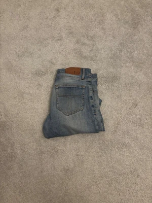 Blå jeans från Tiger of Sweden,W28/L32 - Snygga blå slim fit jeans från Tiger of Sweden med klassisk femficksdesign och diskret slitning framtill. Jeansen har normal midja och raka ben, tillverkade i mjuk denim för en bekväm känsla. Perfekta till en avslappnad stil.