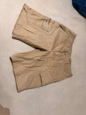 Beige shorts - Shorts för herr. Storlek M. Knappen behöver sys fast igen då den är lös och som synes har en söm lossnat där också. Det är nog inte så svårt att laga men syns heller inte när man har på sig shortsen.