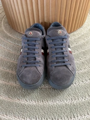 Grå Moncler sneakers i mocka - Snygga sneakers från Moncler i grå mocka med blå snörning och sula. Sidan pryds av Monclers logga samt ränder i vitt, rött och svart. Perfekta för dig som gillar stilrena och exklusiva skor med sportig känsla.