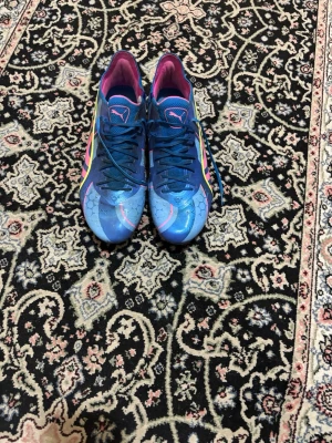 Blåa Puma fotbollsskor med neon - Säljer ett par blåa Puma fotbollsskor med neonrosa och gula detaljer samt hexagonmönster på ovandelen. Skorna har snörning, låg profil och syntetmaterial för bra grepp och kontroll på planen. Insidan är fodrad i rosa och yttersulan har färgglada dobbar.