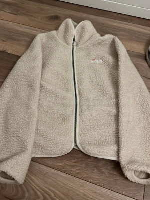 Beige teddyjacka från FILA XS - Mysig beige teddyjacka från FILA i storlek XS. Jackan har dragkedja framtill, hög krage och broderad FILA-logga på bröstet. Tillverkad i fluffigt teddy-material som håller dig varm och ger en trendig look.