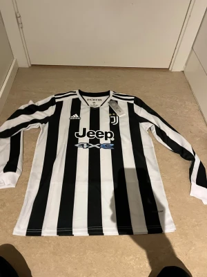 Juventus långärmad matchtröja Adidas - Juventus officiella långärmade matchtröja från Adidas med klassiska svartvita ränder, broderad klubbmärke och tre stjärnor på bröstet. Tillverkad i Primegreen-material och har Jeep som sponsortryck framtill. Perfekt för fotbollsfans och träning.