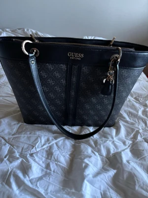 Svart handväska från Guess - Snygg svart handväska från Guess med silverfärgade detaljer och logotyp framtill. Väskan har ett mönster med Guess-loggor över hela ytan, långa axelband och dragkedja upptill. Materialet är konstläder och väskan har en dekorativ nyckelring.