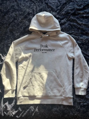 Vit hoodie Peak Performance M - Snygg vit hoodie från Peak Performance med svart logga och text på bröstet. Klassisk känguruficka framtill, ribbade muddar och dragsko i huvan. Tillverkad i mjukt bomullsmaterial som känns skönt mot huden. Perfekt för en avslappnad och stilren look.