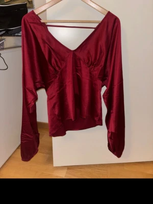 Vinröd satintopp med v-ringning från Nelly  - Helt ny snygg vinröd blus i glansigt satintyg med djup v-ringning både fram och bak. Modellen har långa ärmar och en lös, något kortare passform. Perfekt för dig som vill sticka ut med en elegant och trendig look.