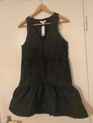 Puffig jaquardklänning - Supersnygg svart kort klänning med v-ringning och glittrig textur. Klänningen är ärmlös och har en volang nedtill som ger en cool och trendig look. Perfekt för dig som vill sticka ut med en elegant men edgy stil.