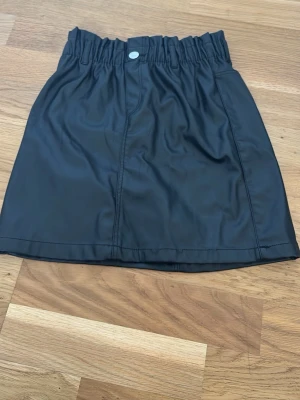 Svart kort kjol med resårmidja - Snygg svart kort kjol med resår i midjan och knappdetalj framtill. Kjolen har en enkel och stilren design, perfekt att matcha med olika toppar. Materialet känns mjukt och har en lätt glansig yta ( det är typ fejk skinkjol )