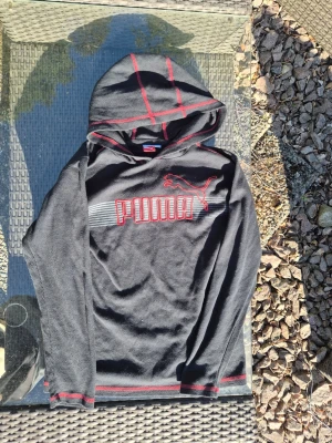 Vintage Svart hoodie från Puma med röd logga - Vintage Svart hoodie från Puma med röd broderad logga och text på bröstet. Hoodien har röda detaljer på huvan och ärmsluten samt en sportig känsla. Tillverkad i ett mjukt och lätt material som passar perfekt till en avslappnad stil. 