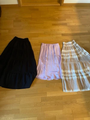 Tre kjolar i olika färger och stilar - Säljer tre snygga kjolar: en lång svart kjol med volanger, en ljuslila kortkjol i slätt tyg och en lång beige och vit kjol med ränder och resår i midjan. Perfekta för att mixa och matcha till olika outfits.