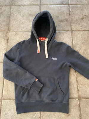 Mörkblå hoodie från Superdry - Snygg mörkblå hoodie från Superdry Orange Label med vit broderad logga på bröstet. Hoodien har huva med vita dragsnören och känguruficka framtill. Mjuk insida och ribbade muddar vid ärmslut och nederkant. Perfekt för en avslappnad stil.