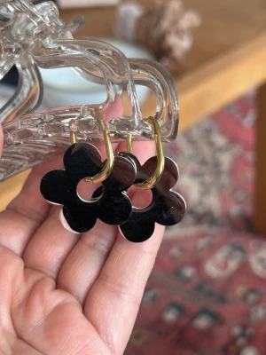 Blommiga örhängen i svart och guld - Snygga örhängen med blommor i svart blankt material och guldfärgade ringar. Blommorna har en hålig mitt och en stilren, modern design. Perfekta för att ge din look en cool och lekfull touch.