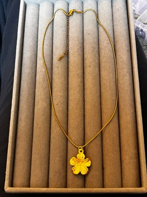 Guldigt halsband med blomma - Snyggt halsband i guldfärg med en blomformad berlock. Kedjan är tunn och elegant, och blomman har fina detaljer som ger ett stilrent intryck. Perfekt accessoar för att lyfta din outfit med en touch av guld och naturinspiration.
