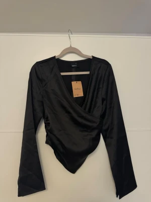 Svart omlottopp från Gina Tricot - Snygg svart omlottopp från Gina Tricot i glansigt satinliknande material. Toppen har lång ärm och v-ringad omlottdesign som ger en trendig look. Perfekt att styla med jeans eller kjol för en cool outfit.❤️