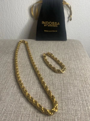 Guldfärgat halsband & armband I - Säljer ett snyggt set med guldfärgat halsband och matchande armband från Indossa by Sweden. Båda har tvinnad kedjedesign och kommer med original svart tygpåse. Perfekt accessoar för att lyfta din outfit.  8mm bred på båda