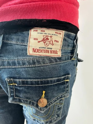 True Religion Joey jeans blå - Säljer ett par blå True Religion Joey jeans med kontrasterande gul söm och snygga detaljer på fickorna. Klassisk femficksmodell med knappar och tydlig logotyp på bakfickan. Materialet är denim och passformen är bootcut, vi har sytt dem lite också så dem blivit kortare som ni kan se på bilden.