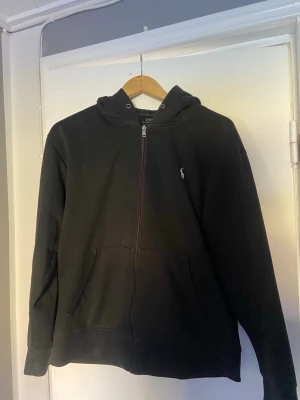 Svart zip hoodie från Polo Ralph Lauren - Säljer en svart hoodie med dragkedja från Polo Ralph Lauren. Klassisk design med huva, fickor framtill och den lilla broderade loggan på bröstet. Perfekt för dig som gillar en clean och stilren look. storleken är m men passar s