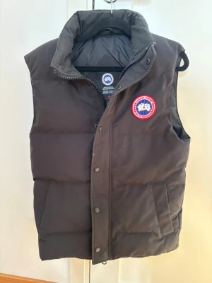 Svart dunväst från Canada Goose - Snygg svart dunväst från Canada Goose med klassisk logotyp på bröstet. Västen har hög krage, dragkedja och tryckknappar framtill samt två fickor. Perfekt för kalla vinterdagar och riktigt varm tack vare dunfyllningen.