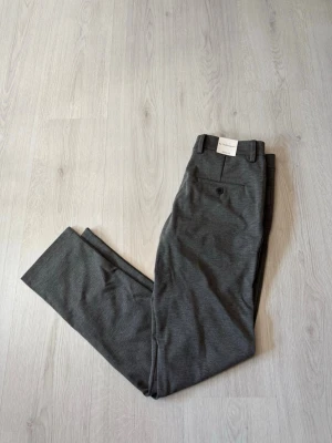 Grå kostymbyxor från TeeShoppen - Snygga grå kostymbyxor från Teeshoppen med klassisk rak passform och bälteshällor. Byxorna har en knappstängning fram och en bakficka med knapp. Perfekta för en stilren look. Materialet är mjukt och har en lätt stretch.