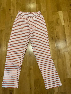 Randiga pyjamasbyxor i vitt och rött stl 34/36 - Pyjamasbyxor i mjukt vitt tyg med röda horisontella ränder och svart texttryck. Byxorna har resår i midjan för extra komfort och en avslappnad passform. Perfekta för mysiga hemmakvällar.