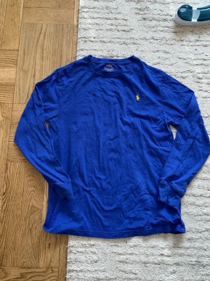 Blå långärmad topp från Polo Ralph Lauren - Klassisk blå långärmad topp från Polo Ralph Lauren med gul broderad logga på bröstet. Tillverkad i mjuk bomull och har en avslappnad passform. Perfekt att styla med jeans eller kjol för en enkel och snygg look.