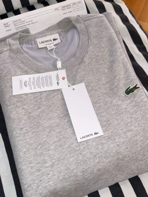 Grå sweatshirt från Lacoste - Klassisk grå sweatshirt från Lacoste med rund halsringning och broderad grön krokodil-logga på bröstet. Tröjan har långa ärmar och ribbade muddar vid ärmslut och nederkant. Perfekt för en avslappnad och stilren look.