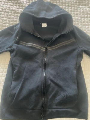 Svart hoodie med dragkedja från Nike - Svart hoodie från Nike med hel dragkedja och huva. Snygga reflekterande ränder över bröstet och ärmen, samt diskret Nike-logga på bröstet. Praktiska fickor framtill och en extra ficka med dragkedja på ärmen. Perfekt för en sportig och avslappnad stil. Storlek S/M