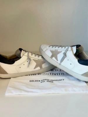 Golden Goose vita sneakers med stjärna - Golden Goose sneakers i vitt läder med grå mockadetaljer och ikonisk stjärna på sidan. Skorna har svarta hälpartier och ljusgrå skosnören. Klassisk låg modell med vintagekänsla och slitna detaljer för en cool streetstil.