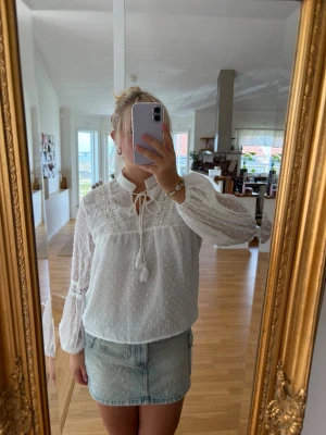 Vit blus med spetsdetaljer - Söt vit blus med broderade spetsdetaljer och knytning vid halsen. Blusen har diskreta prickar i tyget, voluminösa långärmar med spetsinslag och är tillverkad i ett lätt, luftigt material. Perfekt för en trendig och feminin look.