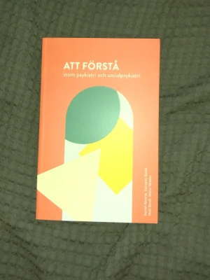 Att förstå inom psykiatri och socialpsykiatri - Kursbok med orange och vit framsida, modern grafisk design och titeln 'Att förstå inom psykiatri och socialpsykiatri'. Perfekt för dig som pluggar eller jobbar inom psykiatri eller socialt arbete. Mjukt omslag och tydlig text.
