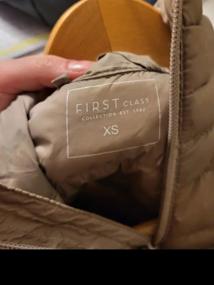 Beige dunväst från First Class XS - Snygg beige dunväst från First Class i storlek XS. Västen har hög krage, dragkedja framtill och två fickor med dragkedja. Quiltad design och lätt vadderad för extra värme. Perfekt för lager-på-lager-stil under kyliga dagar.