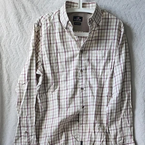 Rutig skjorta från u.s.polo assn  - Klassisk rutig skjorta från u.s.polo assn i vitt med röda och bruna rutor. Skjortan har lång ärm, bröstficka och knappar framtill. Tillverkad i mjuk bomull, perfekt för en avslappnad men ändå stilren look.