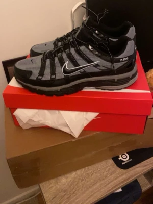 Nike P-6000 svart/grå sneakers - Nike P-6000 sneakers i svart och grått med mesh och syntetiska paneler. Skorna har en sportig vibe med tydliga ränder och en vit Swoosh på sidan. Sulan är robust och ger bra stöd. Perfekta för dig som gillar streetwear och en modern look. Skorna har aldrig använts utanför hemmet och är helt nya. Skorna passar även folk som har storlek 42,5