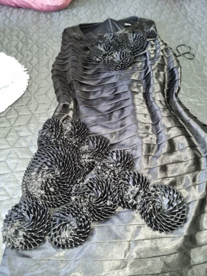 Svart satin klänning med 3D-detaljer - Unik svart klänning i satin med coola 3D-blommor och veckade band över hela plagget. Klänningen har en kroppsnära passform och är ärmlös, perfekt för dig som vill sticka ut med en elegant och modern look. I storlek 42. Oanvänd 