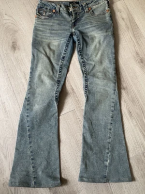 True Religion Joey Low Rise Flare Jeans - Säljer ett par ljusblå jeans från True Religion, modellen Joey Low Rise Flare. Jeansen har låg midja, utsvängda ben och klassiska fem fickor. Snygga slitningar och kontrastsömmar ger en cool vintagekänsla. Perfekta för dig som gillar retrostil! Aldrig använt kostar 1700nya❤️W25, passar xs-s