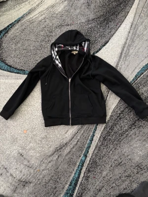 Svart burberry zip hoodie  - En nästan helt ny svart burberry zip hoodie som är använd 1-2 gånger,det finns inga tecken på användning eller något sånt den är helt fräsch.             OBS!! Det är ett fabrikstryck sommar fel i storleken det står XXL men det är till folk som har storlek M-L 
