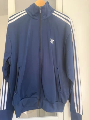 Mörkblå Adidas track jacket - Säljer en mörkblå track jacket från Adidas med klassiska vita ränder längs ärmarna och logotyp på bröstet. Jackan har hög krage, dragkedja framtill. Är i ny skick då Jag använt jackan/tröjan 2-3 gånger 