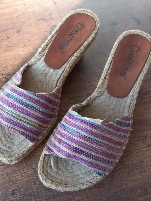 Randiga kilklackssandaler Cinnamon storlek 41  - Färgglada sandaler från Cinnamon med kilklack och öppen tå. Ovandelen har ett randigt tyg i lila, rosa, blått och beige. Sulan är flätad i naturmaterial och innersulan är brun. Perfekta för en avslappnad och trendig sommarlook.