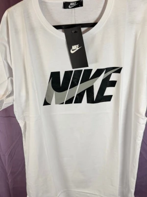 Vit Nike t-shirt med stor logga - Snygg vit t-shirt från Nike med stor svart logga och grå swoosh på bröstet. Klassisk rund halsringning och korta ärmar. Tillverkad i mjuk bomull för skön känsla och stilren look. Perfekt för dig som gillar sportiga och trendiga plagg.