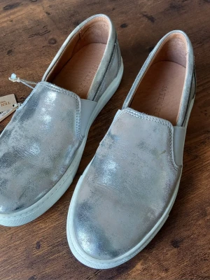 Silvriga loafers från Ten Points storlek 38 - Säljer ett par snygga loafers från Ten Points i metallic-silverfärgat läder. Skorna har en klassisk slip-on design med elastiska paneler på sidorna och vit sula. Perfekta för dig som vill sticka ut med en modern och trendig look.