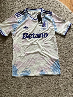 Aston Villa adidas matchtröja - Säljer en Aston Villa matchtröja från adidas i vitt med lila och blå detaljer. Tröjan har klubbmärke, adidas-logga och Betano-tryck på bröstet. Tröjan är i storlek S och är helt ny. Nypris: 1000kr, mitt pris: 299kr. Skriv vid frågor 😊