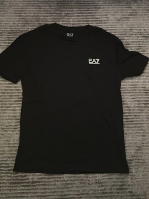 Svart EA7 t-shirt från Emporio Armani - Snygg svart t-shirt från EA7 Emporio Armani med diskret logga på bröstet. Klassisk rund halsringning och korta ärmar. Tillverkad i mjuk bomull som känns skön mot huden. Perfekt till jeans eller shorts för en clean och stilren look.